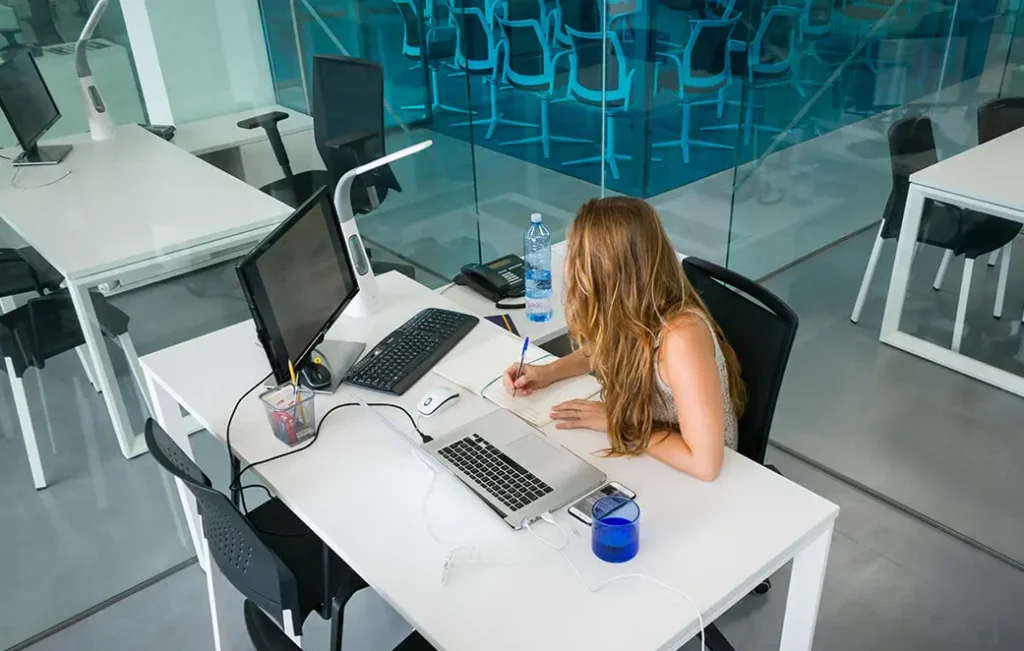 A woman focused on her laptop while seated at a desk in a modern coworking office environment.